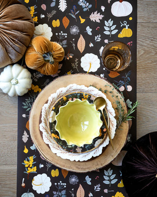 A Harvest Leaves Wreath Shaped Plate from My Mind’s Eye adds a festive touch to a table setting with a green bowl on Thanksgiving dinner plates, accompanied by a spoon and a glass of amber liquid. Decorative gourds and pumpkins are stylishly arranged on a floral-patterned table runner.