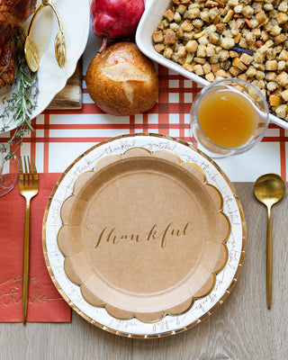 An elegant Thanksgiving celebration ambiance is created with a table setting featuring a "Harvest Scallop Kraft Plate" by My Mind’s Eye, complemented by gold foil accents, a stuffing dish, utensils, a red napkin, bread, pomegranate, and a glass of juice on a red and white checkered tablecloth.