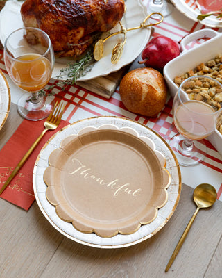 A Thanksgiving table features a Harvest Scallop Kraft Plate by My Mind’s Eye amidst the feast, including a roast turkey, stuffing, pomegranate, and bread rolls. Drinks in stemmed glasses shimmer next to gold-colored cutlery, all creating a festive scene enhanced by elegant party plates with gold foil accents.