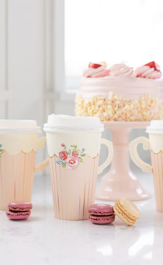 Tea Party Handled Paper Cups by My Mind’s Eye, featuring floral designs, sit beside pink and cream macarons. In the background, a cake elegantly decorated with pink frosting is displayed on a stylish stand.