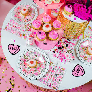 A festive table setup features pink and white cupcakes, decorated plates, Valentine's Day Love Notes Napkins by Daydream Society, cups, and gold foil accents on a pink surface adorned with confetti.