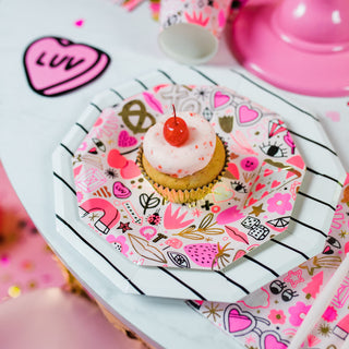 A cupcake with white frosting and a cherry on top sits on a Love Notes Plate by Daydream Society, featuring gold foil-pressed geometric patterns. It's surrounded by pink decorations and a heart-shaped cutout with "luv" for Valentine's Day celebrations.