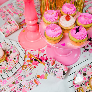 A table setup filled with pink-themed party decorations showcases cupcakes with pink frosting and disposable "Love" cups. Gold foil accents add elegance, while confetti is scattered among the playful Daydream Society's Love Notes Plates for Valentine’s Day.