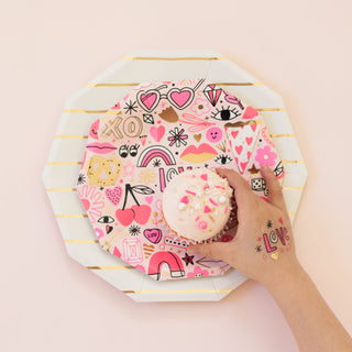 A hand reaches for a pink frosted cupcake on a Daydream Society's Love Notes Plate, vibrant with doodles and hearts. Neon colors enhance the design, complemented by a playful temporary "love" tattoo on the wrist.