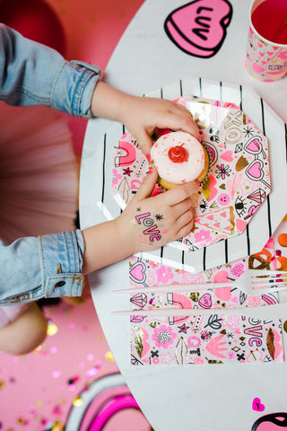 A child holds a cupcake with heart decorations, surrounded by neon colors and confetti on a table. Daydream Society’s Love Notes Plates with gold foil elements add charm to the festive Valentine’s scene.