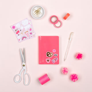 A pink notebook with Valentine's stickers, a pen, scissors, washi tapes, pom-poms, and a sheet of small stickers from the Daydream Society love notes sticker set lies on a light pink surface.