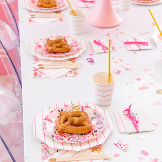 The party table features Love Notes Plates by Daydream Society with pretzels, striped cups, napkins, and vibrant decorations on a white table with gold foil-pressed elements.