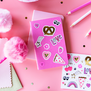 The "Love Notes" sticker set by Daydream Society features a pink notebook decorated with stickers like a pretzel, pizza slice, and "Love," all on premium matte paper. Valentine's Day stickers enhance its charm, surrounded by fluffy pens and pink stationery on a polka dot surface for whimsy.