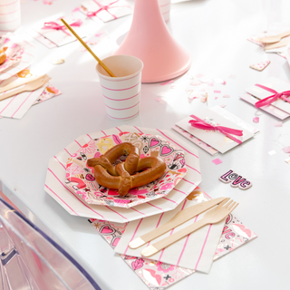 A pretzel rests on a pink-themed table with Daydream Society's Love Notes Plates, a cup, wooden cutlery, and gold foil elements. Confetti adds festivity alongside a celebratory party hat.