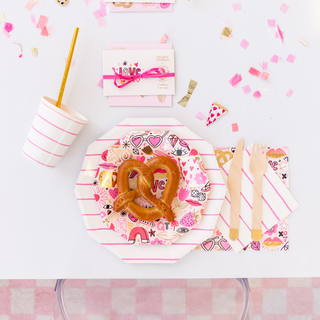 A Valentine's Day table setting features pink and white striped plates, heart-shaped waffles, a striped cup with a straw, wooden cutlery, Love Notes Napkins by Daydream Society, and gold foil confetti with an envelope reading "Love" scattered around.