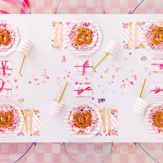 A tabletop features Daydream Society's decorative pink and white Love Notes Plates, pretzels, cups with straws, and wrapped gifts. Gold foil-pressed elements shimmer with confetti as "LOVE" letters are scattered across the surface.