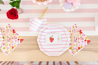 A table setting with striped pink plates, wooden cutlery, and a gold foil-pressed cup with a gold straw. Heartbeat Gang Large Napkins by Daydream Society complement the strawberry card. The scene is completed with macarons and a red rose in a vase, radiating charm.