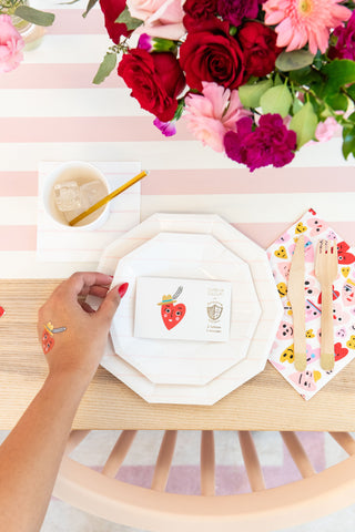 A person with a strawberry sticker holds an envelope above a neon-accented table set with a drink, wooden utensils, floral decorations, and festive Valentine's Day ambiance, featuring Heartbeat Gang Large Napkins by Daydream Society.