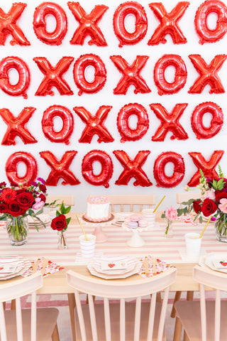 A festive table features a cake and vibrant flowers, with red "XOXO" balloons adding charm to the backdrop. Neon and gold Heartbeat Gang Small Plates by Daydream Society elevate the scene with modern elegance.