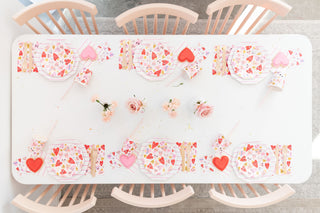 The table is ready for a party with pink and white plates, Daydream Society's Heartbeat Gang Large Napkins featuring heart motifs, gold foil-pressed cups beside neon straws, and pink roses at the center. Six chairs complete this charming scene.