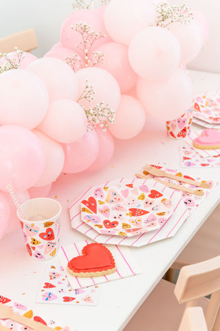 A decorated table showcases Valentine's Day with Daydream Society's Heartbeat Gang Small Plates, neon and gold foil designs, pink balloons, heart-themed napkins, a paper cup with a straw, and a heart-shaped cookie on a white surface.