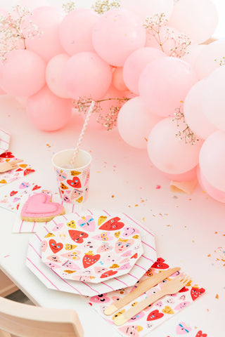 A festive table setting with Heartbeat Gang Small Plates from Daydream Society, a wooden cutlery set, a pink iced cookie, and a pink balloon garland.