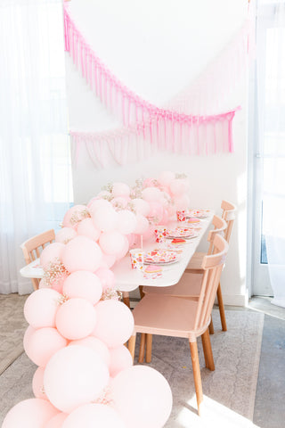 A dining table is adorned with pink balloons and Heartbeat Gang Small Plates by Daydream Society. Neon and gold foil-pressed streamers hang on the wall in the background, creating a festive atmosphere.