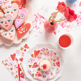 A table set with Daydream Society's Heartbeat Gang Small Plates showcases heart-themed cookies, a radiant cupcake, a vibrant red drink, and a pink vase filled with lush red flowers. Neon and gold foil-pressed details add charm alongside heart-patterned napkins.