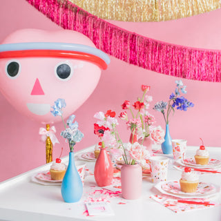 A festive table features floral arrangements in pastel vases, gold foil-pressed details, cupcakes, and vibrant decorations like a large balloon and neon accents set against a pink backdrop. Heartbeat Gang Large Napkins by Daydream Society add an extra touch of love to the celebration.