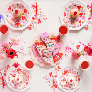 A vibrant Valentine's Day-themed table setting features heart-shaped cookies, floral decorations, and red drinks on a white table adorned with Daydream Society's Heartbeat Gang Large Napkins decorated in gold foil and neon accents.