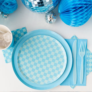 A Check It! Out of the Blue Dessert Plate from Jollity & Co is paired with matching cups, checkered napkins, and plastic cutlery, set among blue and silver disco balls for a complete party setup.