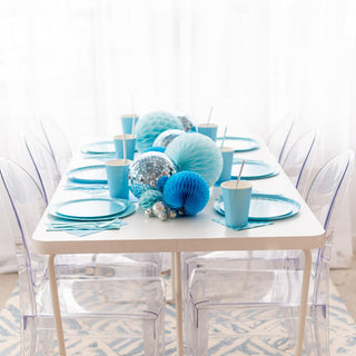 A white table is adorned with Check It! Out of the Blue Dessert Plates by Jollity & Co, matching cups, checkered napkins, and blue-silver decorative spheres as centerpieces. Clear chairs surround the setup.