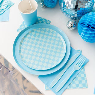 Enhance your party checklist with Jollity & Co's Check It! Out of the Blue Dessert Plates. Create a charming setting with blue checkered plates, matching napkins, a blue paper cup, and plastic cutlery paired with decorations like a disco ball on a white surface.