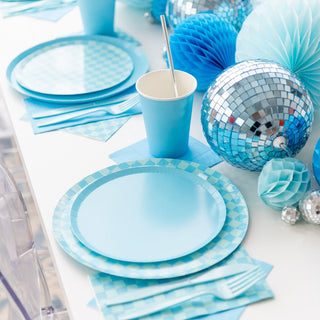 The table is set with Jollity & Co's Check It! Out of the Blue Dessert Plates, checkered print napkins, and a paper cup. Silver disco balls and blue decorative balls are artfully arranged, completing the perfect party checklist.