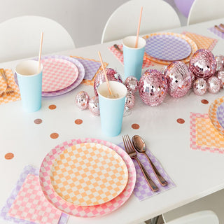 A party table includes Jollity & Co's Check It! Purple Posse Dinner Plates with matching print napkins, blue cups with straws, and silverware. Pink disco balls and confetti centerpieces add a festive touch to the celebration.