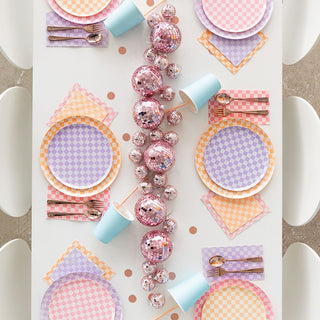 A table setting featuring Jollity & Co's Check It! Purple Posse Dinner Plates with two-tone pastel checkered print, napkins, and blue cups. A centerpiece of pink disco balls runs down the middle, surrounded by silverware.