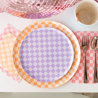 Three Jollity & Co Check It! Purple Posse Dinner Plates with purple and orange checkered patterns are stacked on a table with matching checkered print napkins. A cup with ice and utensils sit nearby.