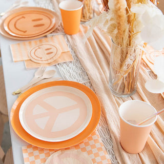 The table embraces retro-chic style with orange Jollity & Co Peace & Love Peace Dessert Plates, cups, and napkins featuring peace symbols and smiley faces. A neutral runner complements a tall vase of dried flowers, while daisy accents subtly enhance the theme of peace and love.