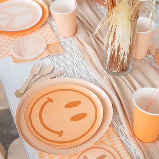 A whimsical table set with Jollity & Co Peace & Love Smile Dessert Plates, cups, and wooden utensils rests on a checkered ivory lace tablecloth. Pampas grass in a vase adds charm as the centerpiece.