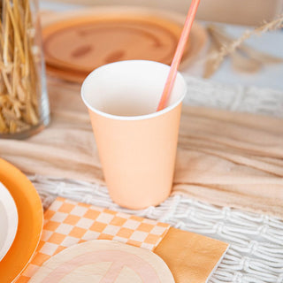 A peach paper cup with an orange straw and Jollity & Co's Peace & Love Smile Dessert Plates create a whimsical, retro-chic vibe on a table accented by a plaid napkin. Dried wheat stalks in the background enhance the nostalgic charm, invoking tranquility.