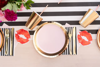 A table setting featuring Jollity & Co's eco-friendly Petal Shade metallic plates, cups, and cutlery on a black-and-white striped tablecloth. Red lipstick print napkins add a playful touch, while pink flowers in the corner enhance the ambiance of the Shades Collection.