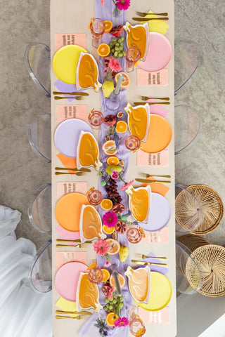 A table setting features Jollity & Co's vibrant Petal Shade Petal Dinner Plates, golden cutlery, assorted fruits, and flowers artfully laid out on a purple runner.