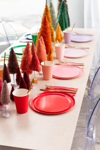 The festive table gleams with Jollity & Co's vibrant Petal Shade Dinner Plates, colorful cups, and gold utensils. Miniature decorative trees charm the center of the table, adding whimsy.