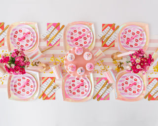 Aerial view of a pink-themed party table featuring Jollity & Co's Candy Striped Dinner Plates alongside floral arrangements and cupcakes on a cake stand. Striped napkins and gold utensils complete the setting.