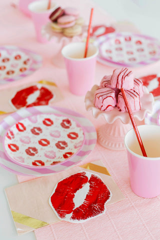 A pink-themed party table features Jollity & Co's Candy Striped Dinner Plates, napkins with red lip prints, and cups with straws. Macarons and marshmallows elegantly rest on a cake stand, completing the festive spread.