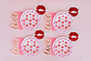 Four Candy Striped Dinner Plates by Jollity & Co, napkins, and "love" cutouts form an enchanting pink-themed party setting on a soothing pink background.