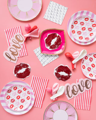 A pink-themed party scene unfolds with Jollity & Co's Candy Striped Dinner Plates and napkins adorned with lip prints. "Love" cutouts nestle among deflated balloons on a vibrant pink background, creating a whimsical and festive atmosphere.