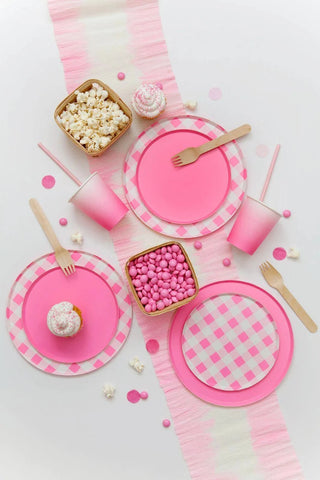 A neatly arranged, playful pink-themed party table setting featuring Oh Happy Day Neon Rose Pattern Plates - 7 inch, wooden utensils, cups of popcorn and sweets, and confetti, all atop a white surface.
