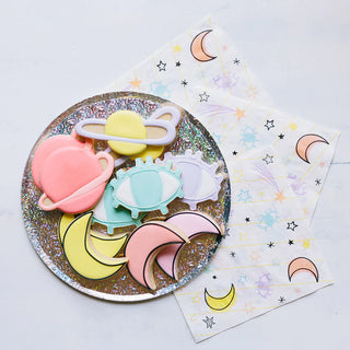 A collection of colorful, celestial-themed silicone teethers and coordinating napkins featuring moon and star patterns, all displayed on a glittery round Party Plates designed for special occasions by Loop by Frankie.