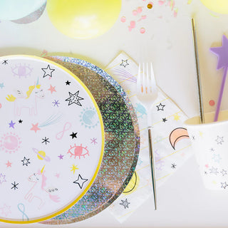 A festive table setting featuring Loop by Frankie's iridescent paper plates, a glittery round placemat, a golden fork, a party cup adorned with stars, confetti, and a star-topped wand, suggesting