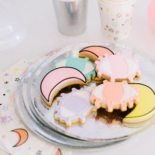 A collection of pastel-colored, space-themed cookies artfully arranged on Loop by Frankie's Iridescent Paper Plates, complemented by a festive background with scattered star decorations and party cups.