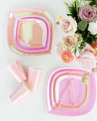 Posh Just Peachy Cups by Jollity & Co, featuring iridescent pink plates and gold foil, are displayed next to a bouquet of roses and ranunculus on a white surface.