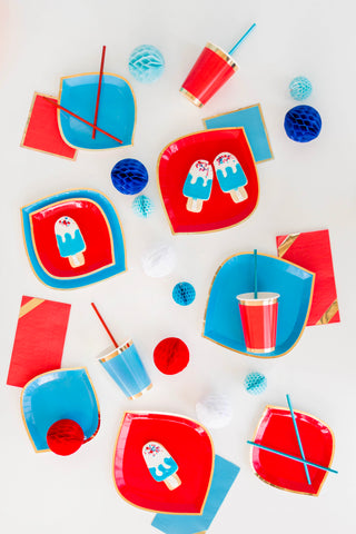 Vibrant tableware includes red and blue plates with ice pop designs, Jollity & Co's Posh Ruby Kiss Cups featuring straws and gold foil accents, and decorative paper balls on a pristine white surface.