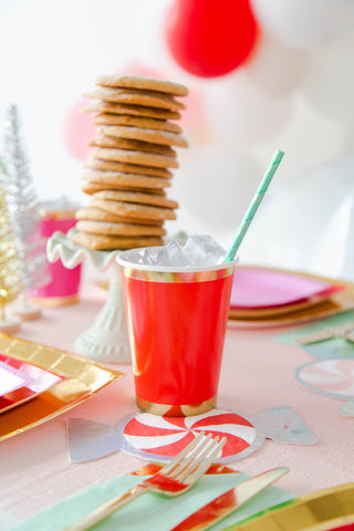 A Posh Ruby Kiss Cup by Jollity & Co, filled with ice and a straw, sits on a table next to cookies and colorful plates from the Luxe Collection. Balloons and festive decorations in the background enhance the celebratory atmosphere.