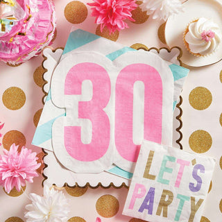 A colorful 30th birthday celebration setup featuring a bold, durable Slant Jumbo Shaped Napkin "30" centerpiece, a "let's party" bag, cupcakes, and elegant pink flowers, all arranged on